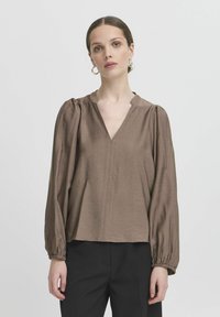Blusa marrone a maniche lunghe con scollatura a V, spalle arricciate e polsini elastici. Realizzata in tessuto testurizzato, presenta una cucitura sottile.