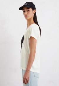 Witte t-shirt met korte mouwen en een zwart stergrafiek aan de voorkant; gecombineerd met lichtblauwe jeans; gedragen met een zwarte pet.
