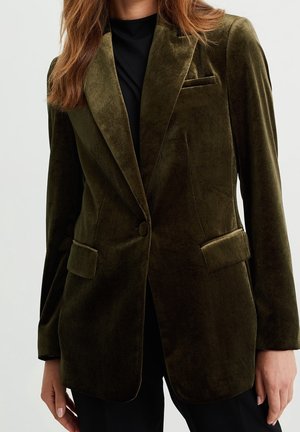 Veste en velours vert olive avec revers crantés, fermeture à un bouton et poches à rabat portée sur un haut noir.