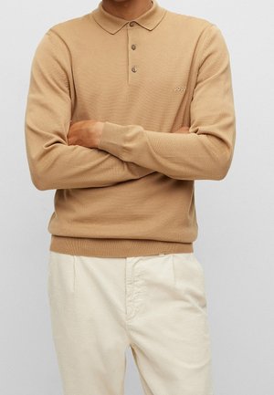 Person iført en beige langærmet polo sweater med "BOSS" logo og lyse cremefarvede bukser, arme krydset, mod en ensfarvet grå baggrund.
