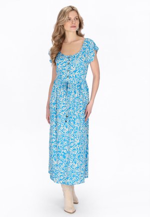 Kobieta z długimi falującymi włosami, ubrana w niebieską midi sukienkę w wzory z guzikami i paskiem w talii, w połączeniu z beżowymi botkami za kostkę.