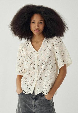 Žena s objemnými kučeravými vlasmi oblečená v bielej blúzke so zapínaním na gombíky a sivých džínsoch, stojaca s rukami v vreckách pred jednoduchým pozadím.