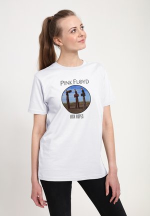 Kvinde med brunt hestehale iført hvid Pink Floyd "High Hopes" t-shirt med tre mænd på stylter mod en blå himmel.