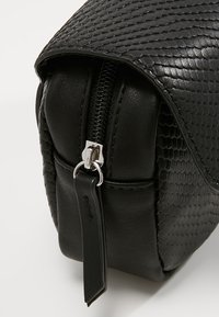 Sac en cuir synthétique noir avec une texture peau de serpent, fermeture éclair en métal argenté et rabat supérieur plat. Présente une petite étiquette en cuir et une forme arrondie.