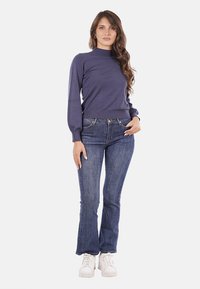 Maglione blu navy con collo alto, maniche lunghe, abbinato a jeans in denim svasati. Il modello indossa sneaker bianche. Design semplice e aderente.