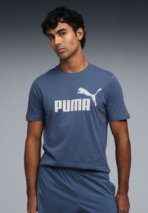 Mėlynas medvilninis marškinėlis su dideliu baltu "PUMA" logotipu ir šuoliuojančio puma grafika priekyje. Trumpomis rankovėmis ir apvaliu kaklu.