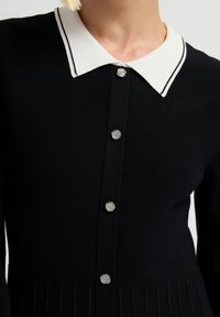 Abito nero a maglia con colletto bianco decorato da due strisce nere. Sei bottoni a forma di fiore in argento e dettagli a coste verticali sull'orlo.