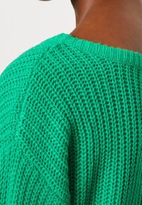 Grön stickad tröja med ett löst stickat mönster och ribbad halsringning, som framhäver textur och en klar färg.