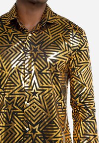 Sort og guld mønstret skjorte med geometriske stjernedesigns og striber. Glat, skinnende stof med knapper ned ad fronten.