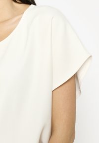 Blusa beige chiaro con vestibilità rilassata, tessuto morbido, scollo rotondo e maniche corte con dettagli di pieghe sottili.
