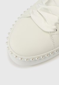 Λευκό δάχτυλο αθλητικού παπουτσιού με κορδόνια από σατέν κορδέλα και πέρλες κατά μήκος της άκρης της σόλας σε απλό λευκό φόντο.