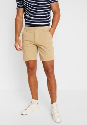 Mann trägt beige Shorts, navy-weiß gestreiftes T-Shirt und weiße High-Top-Sneaker, steht mit einer Hand in der Tasche.