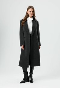 Grijze wollen jas met een lang, getailleerd ontwerp, dubbele knopenrij, moderne revers en een hoogsluitende hals, gedragen over zwarte aangesloten broek en laarzen.