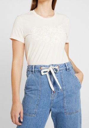 Mujer que lleva una camiseta blanca de manga corta con un bordado floral sutil y jeans azules de talle alto atados con un cinturón blanco de cordón.