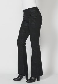 Pantalones negros con estampado floral, ajustados, con patas acampanadas, bolsillos laterales y cierre de cremallera. Combinados con botas de tacón alto negras.