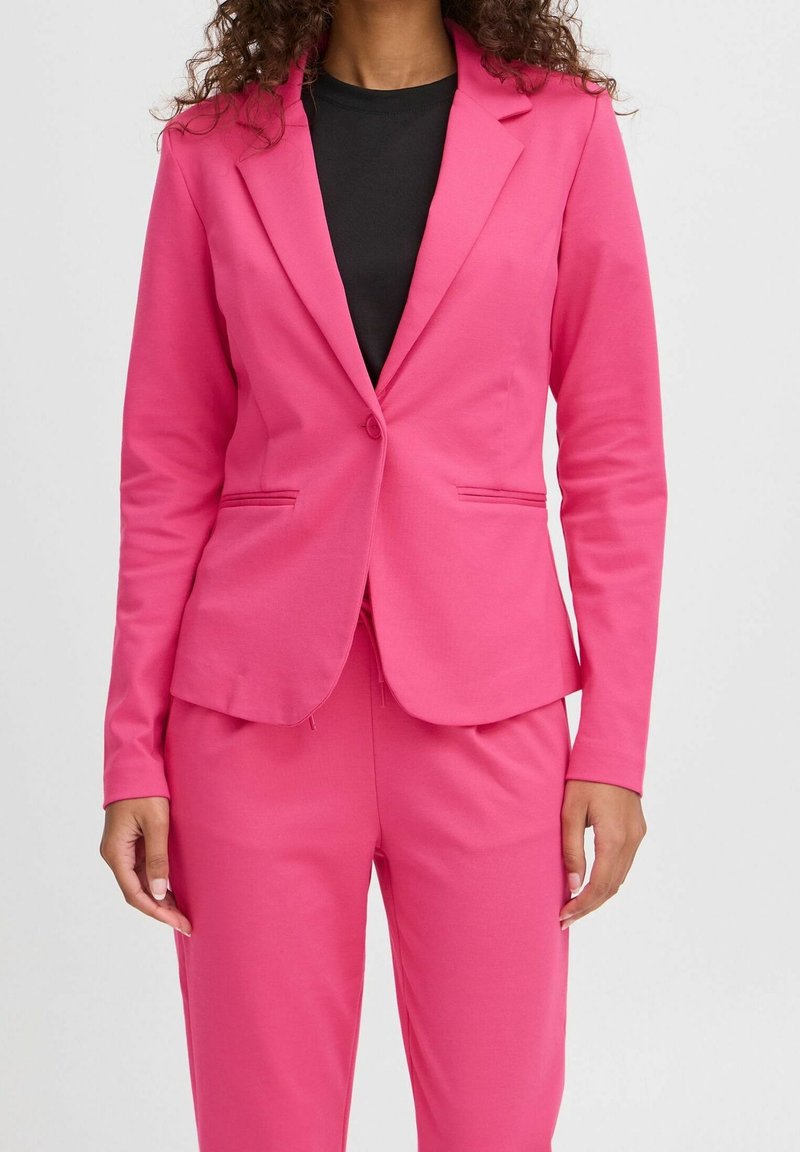 Femme portant un blazer rose vif et un pantalon assorti sur un top noir, sur un fond clair uni.