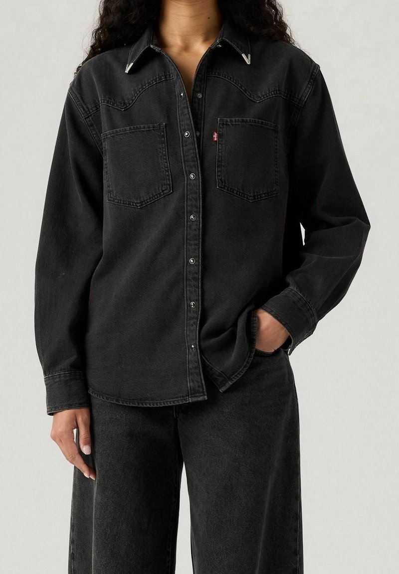 Femme portant une chemise en denim noire boutonnée avec deux poches poitrine et un pantalon en denim noir assorti, une main dans la poche.