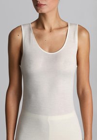 Donna che indossa un top bianco senza maniche con scollatura a girocollo, mostrata dalle spalle ai fianchi contro uno sfondo grigio uniforme.