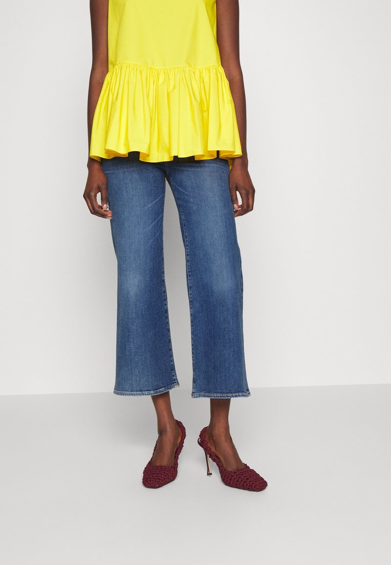 Femme portant un jean court bleu, un haut jaune à volants et des escarpins bordeaux à bout pointu avec une texture tissée.
