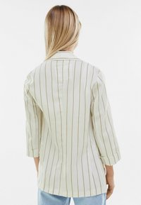 Světle béžová pruhovaná košile s tříčtvrtečními rukávy, která má zadní rozparek a měkkou texturu látky, zobrazená z pohledu zezadu.