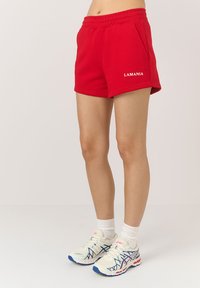 Czerwone spodenki sportowe z elastycznym pasem i kieszeniami bocznymi, z logo "LAMANIA" w kolorze białym. Nosi się je z białymi skarpetkami i jasnymi butami sportowymi.