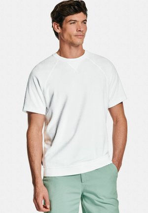Homme aux cheveux bruns portant une chemise blanche à manches courtes et un pantalon vert clair, se tenant avec une main dans sa poche, regardant sur le côté.