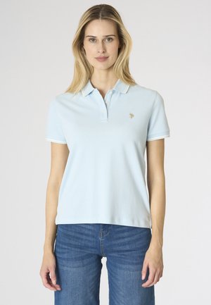 Donna che indossa una polo azzurro chiaro a maniche corte con colletto e jeans blu, in piedi davanti a uno sfondo bianco uniforme.
