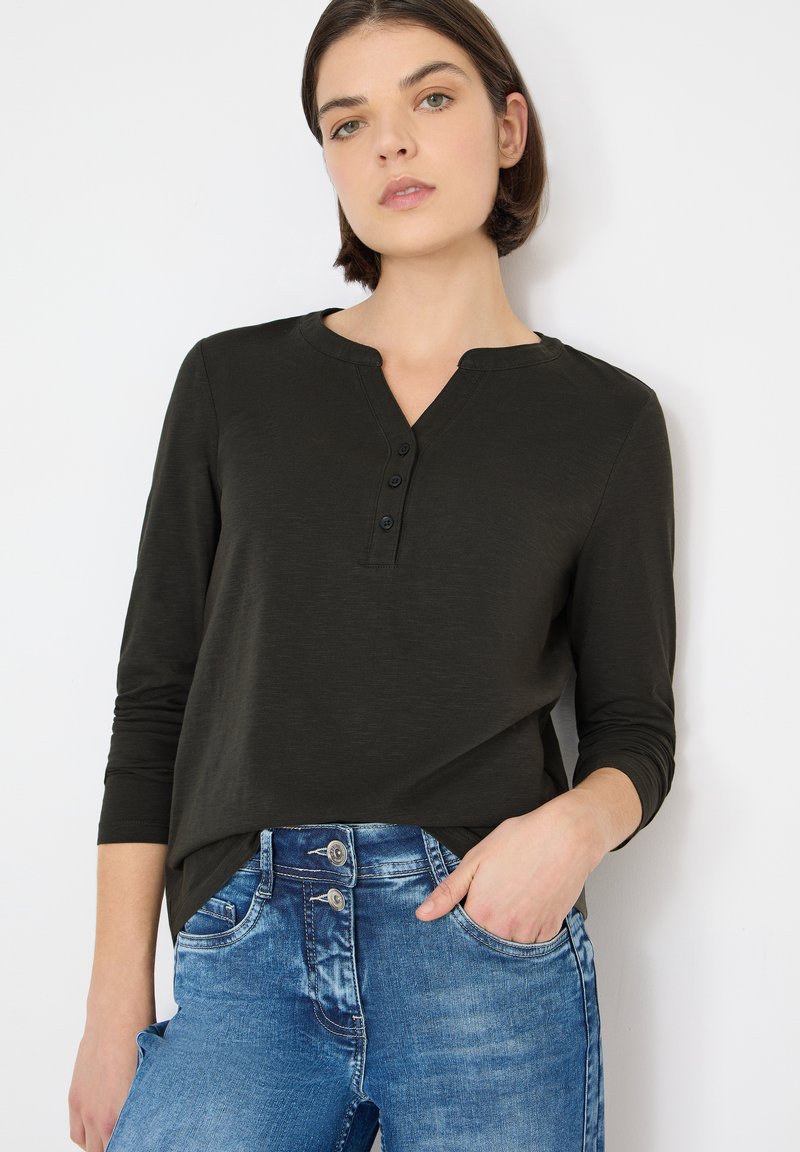 Maglietta verde scuro a maniche lunghe con scollo henley e tre bottoni. Indossata con jeans di denim blu con due bottoni sulla vita.