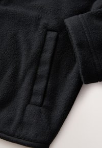 Giacca in pile nera con una texture morbida, dotata di una tasca laterale e maniche con polsini per maggiore comfort e funzionalità.