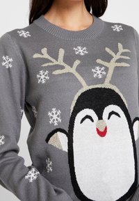 Harmaa villapaita, jossa on pingviinimuotoilu, kultaiset sarvet, valkoisia lumihiutaleita ja teksturoitua neulekuosia. Koristeellinen kuvio on keskiössä edessä.