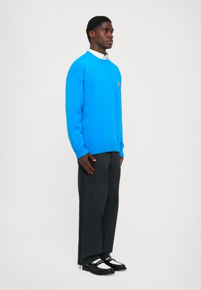 Blauer Strickpullover mit gerippten Bündchen und Saum, kombiniert mit dunklen Hosen und schwarzen Slippern mit goldener Kettenakzent.