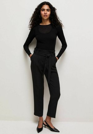 Vrouw met krullend haar, gekleed in een zwarte top met lange mouwen, hooggetailleerde paperbag-broek met riem en zwarte slingback hakken, staand tegen een effen muur.