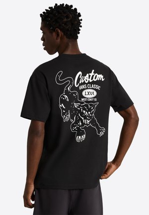 Osoba z krótkimi kręconymi włosami ubrana w czarną koszulkę z białą grafiką warczącej pantery i napisem „Custom Vans Classic LXVI West Coast Co.”