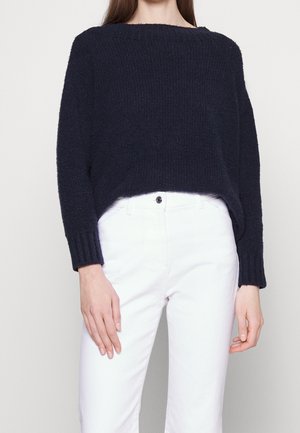 Personne portant un pull texturé bleu marine foncé à manches longues et un pantalon blanc taille haute sur un fond uni.