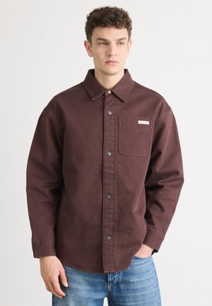 Camicia button-up bordeaux con taschino sul petto, realizzata in tessuto resistente. Caratterizzata da un colletto aperto e bottoni grigi, indossata sopra jeans blu.