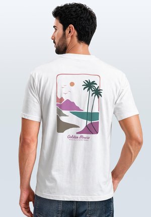 Mann mit dunklem Haar und Bart, der ein weißes T-Shirt mit buntem Küstendesign mit Palmen, Sonne, Vögeln und Bergen trägt.
