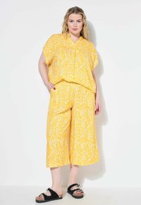 Gelbes Zweiteiler-Outfit mit einem kurzärmeligen Oberteil und weit geschnittenen Hosen mit einem weißen abstrakten Muster; schwarze Plateausandalen.
