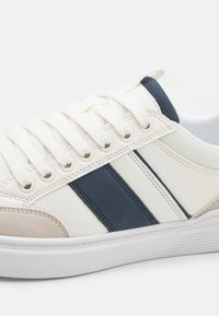 Weiße Sneakers mit strukturiertem Lederoberteil, marineblauen und beigen Streifen, atmungsaktiven Mesh-Elementen und Gummisohle mit dezenten Rillenmustern.