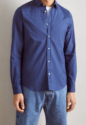 Mann trägt ein marineblaues Hemd über einem weißen Unterhemd, kombiniert mit hellblauen Jeans, steht vor einem schlichten Hintergrund.