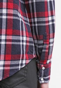Chemise à carreaux rouge, bleu marine et blanc à manches longues, poignets avec boutons blancs, portée sur un pantalon noir avec des coutures visibles.