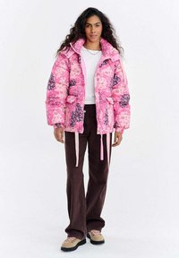 Różowa pikowana kurtka z kwiatowymi wzorami i różowymi troczkami, w połączeniu z brązowymi spodniami dzwonami i beżowymi butami. Projekt ma wysoki kołnierz.