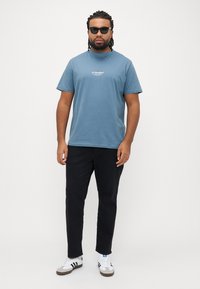 Blå bomulds t-shirt med korte ærmer og hvidt logo, kombineret med sorte bukser og hvide sneakers med sorte striber.