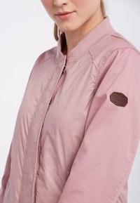 Veste bomber rose avec un devant matelassé, un col côtelé et un patch en cuir marron sur la manche. Texture lisse et coupe ajustée.