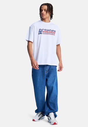 Wit T-shirt met korte mouwen met een kleurrijk grafisch logo, gecombineerd met losse blauwe spijkerbroeken en witte sneakers met rode accenten.