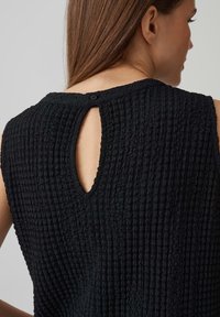 Top senza maniche nero in tessuto testurizzato con chiusura a chiave sul retro e motivo a coste, dotato di un bottone sul collo per un dettaglio aggiuntivo.