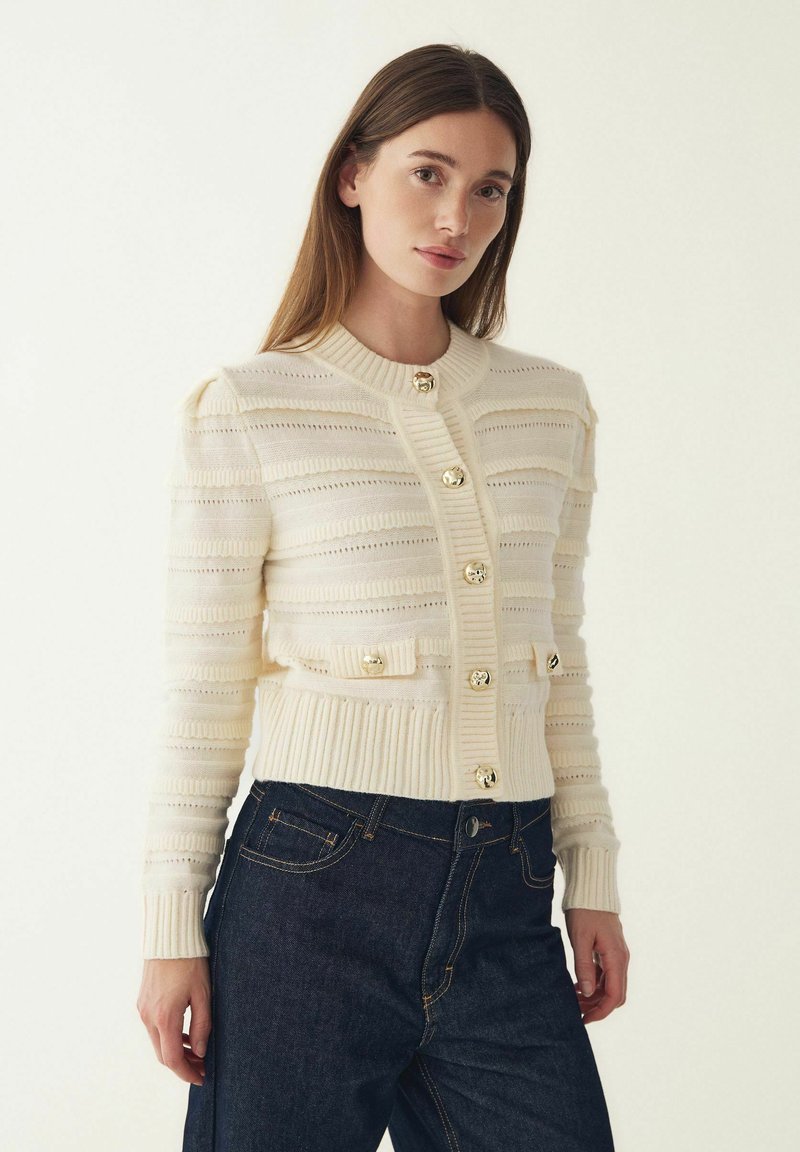 Kardigan in Cremetönen mit gerippter Struktur, horizontalen Streifen und sechs goldenen Knöpfen. Verfügt über einen Rundhalsausschnitt und eine taillierte Passform.