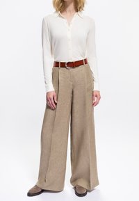 Donna che indossa una camicia cream a maniche lunghe con bottoni infilata in pantaloni beige a gamba larga, con una cintura marrone e scarpe taupe.