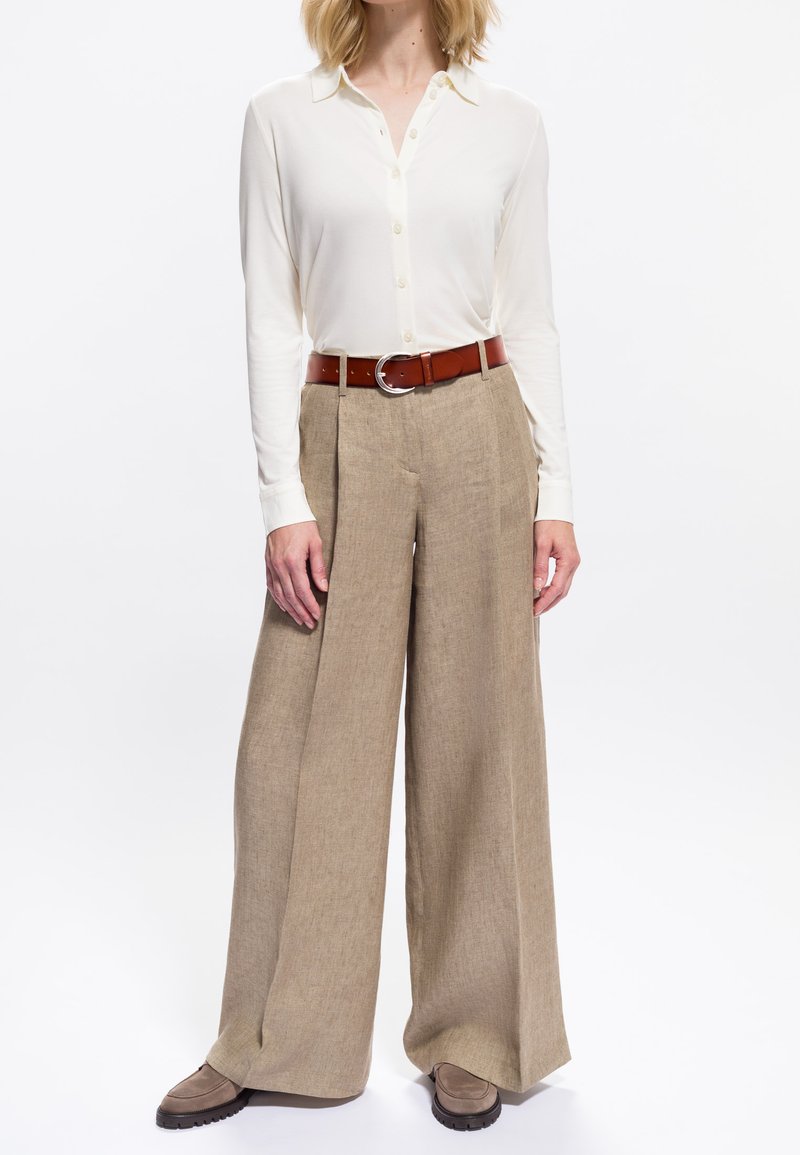 Donna che indossa una camicia cream a maniche lunghe con bottoni infilata in pantaloni beige a gamba larga, con una cintura marrone e scarpe taupe.