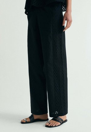 Mujer vistiendo pantalones negros de pierna ancha con patrones recortados en los laterales y sandalias planas negras con detalles de hebilla plateada.