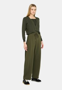 Maglione verde oliva a scollo a V con maniche lunghe, indossato sopra una maglietta nera con motivo. Abbinato a pantaloni larghi verde oliva e sneakers nere.
